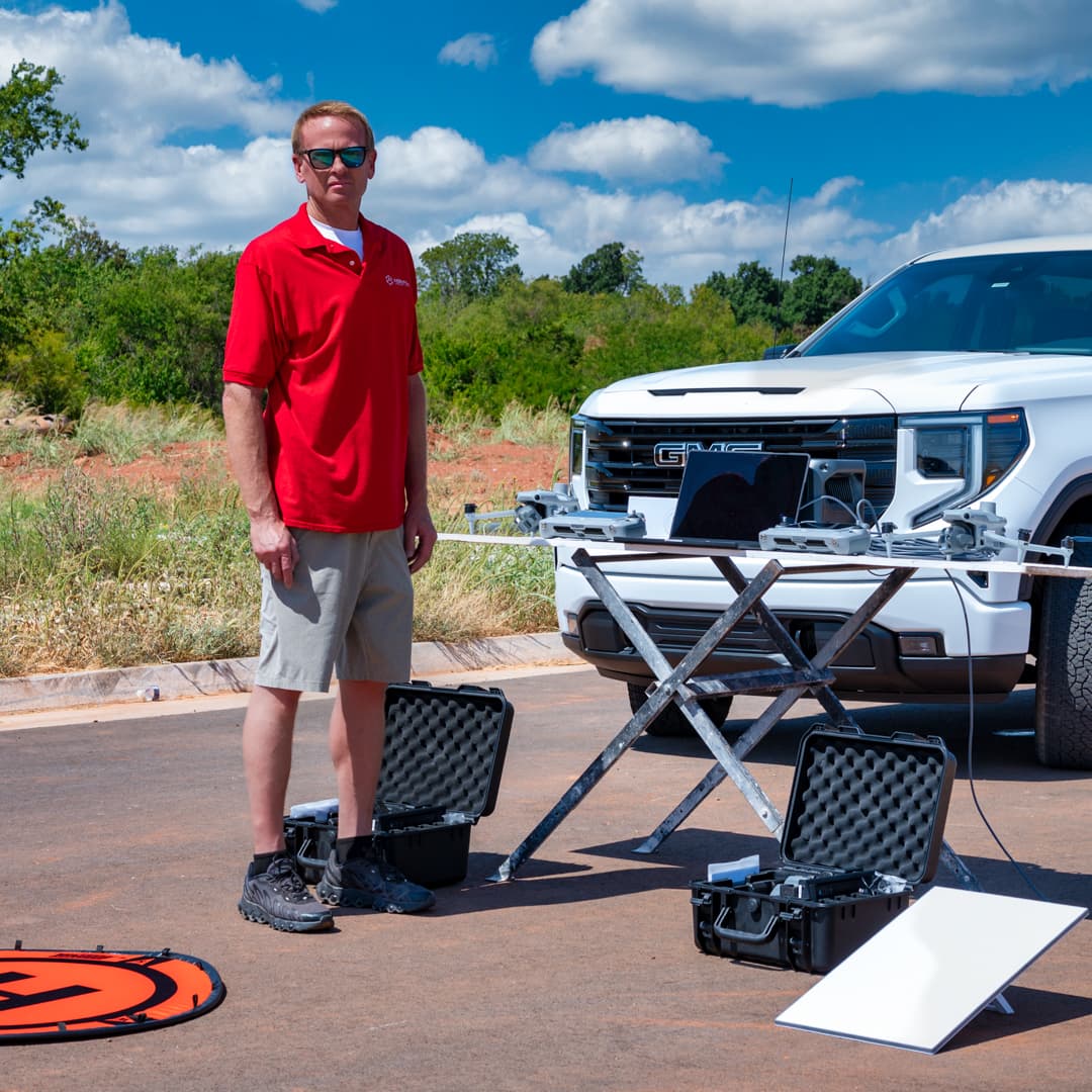 Aerial Perception pilot on location beside a field table of drones, controllers and a satellite uplink, with a white pickup truck behind